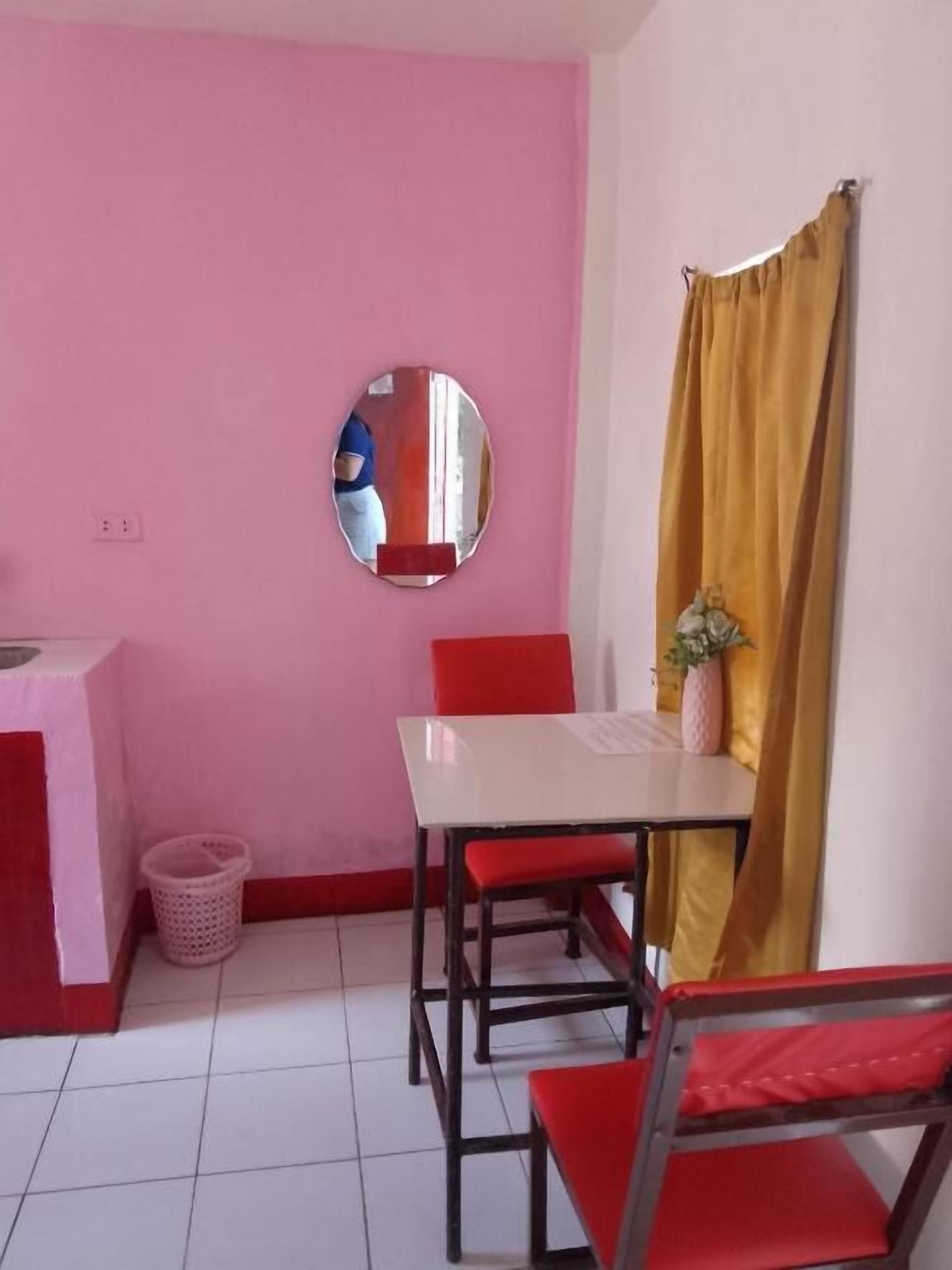Una stanza piccola con pareti rosa, un tavolo bianco e sedie rosse. Si nota uno specchio al muro, una tenda gialla e una piccola pianta sul tavolo.