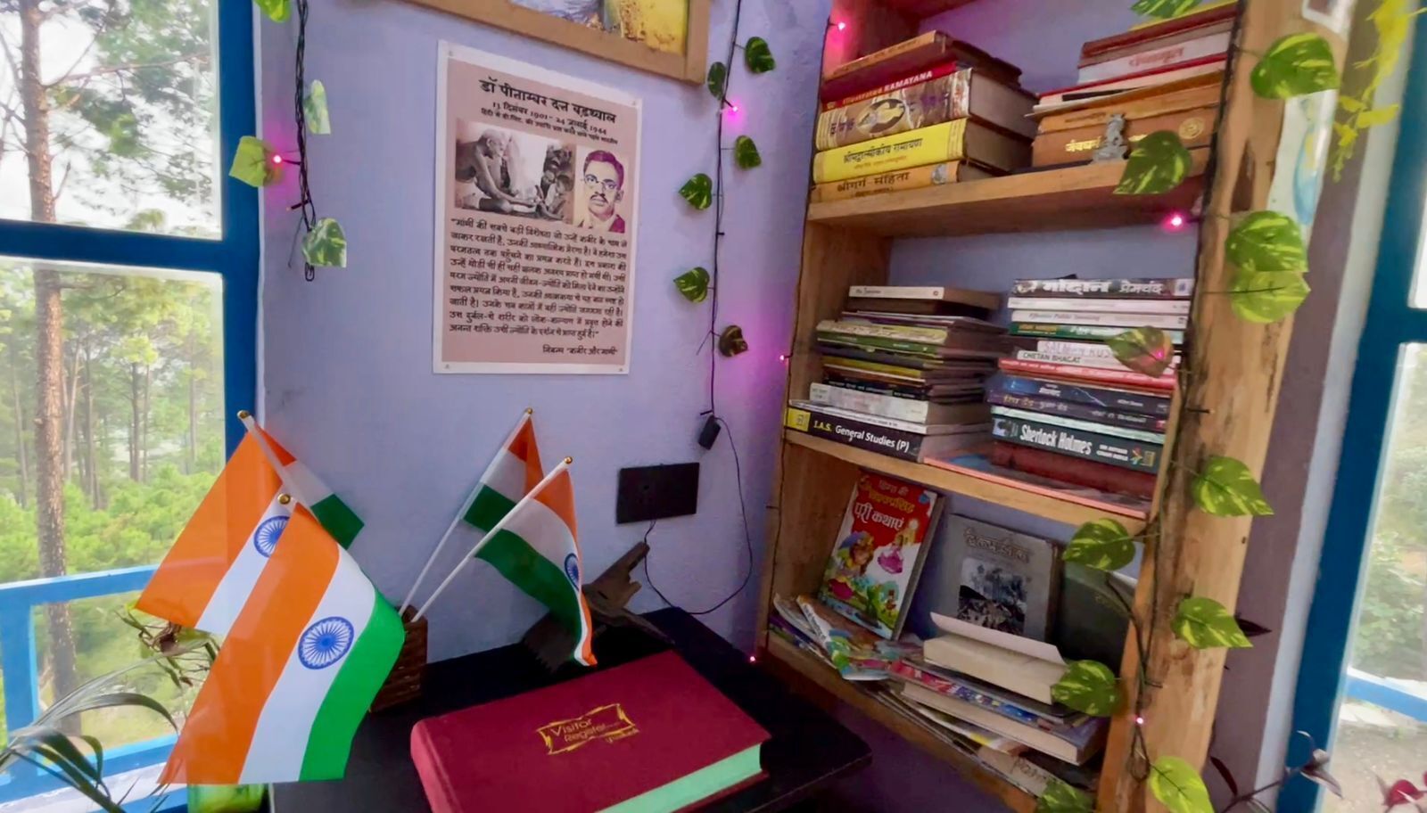 A room with a bookshelf, two Indian flags, a framed poster, and a window with a view of trees.