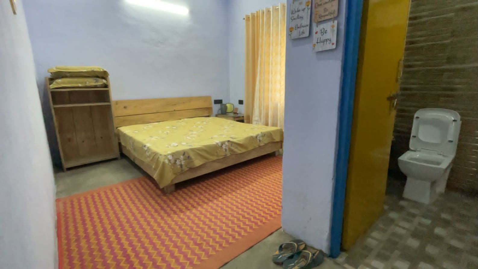 A small, simple room with a bed, a wardrobe, a toilet, and a small table with a chair.