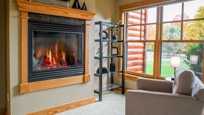 A cozy living room with a fireplace, a glass shelf unit, and a view of a wooden cabin through large windows.