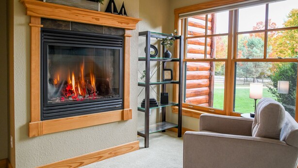 Un salon chaleureux avec une cheminée, une étagère en verre et une vue sur un chalet en bois, visible à travers de grandes fenêtres.