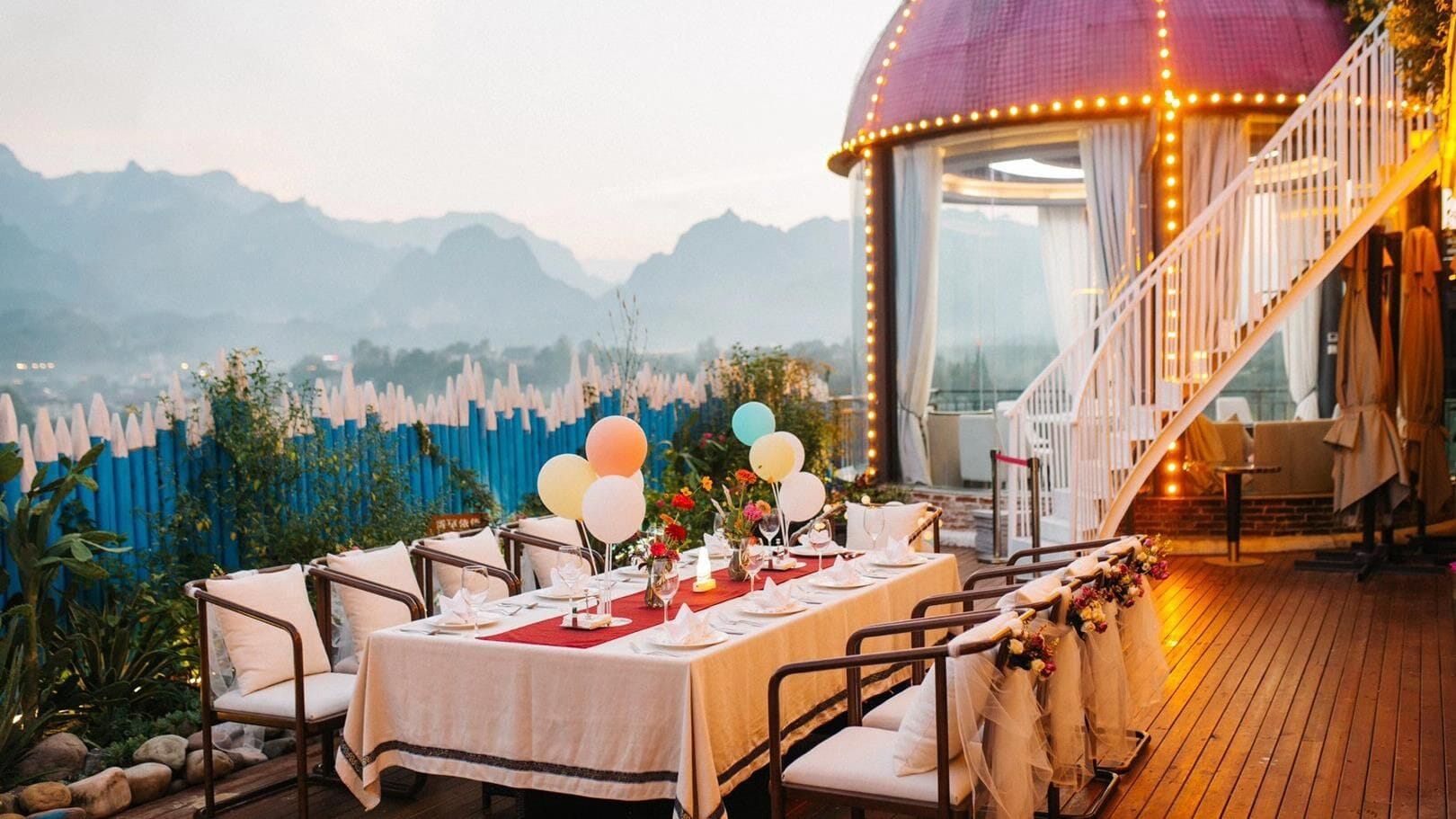 An outdoor dining area with a table set for a meal, surrounded by chairs, and a view of mountains in the background.