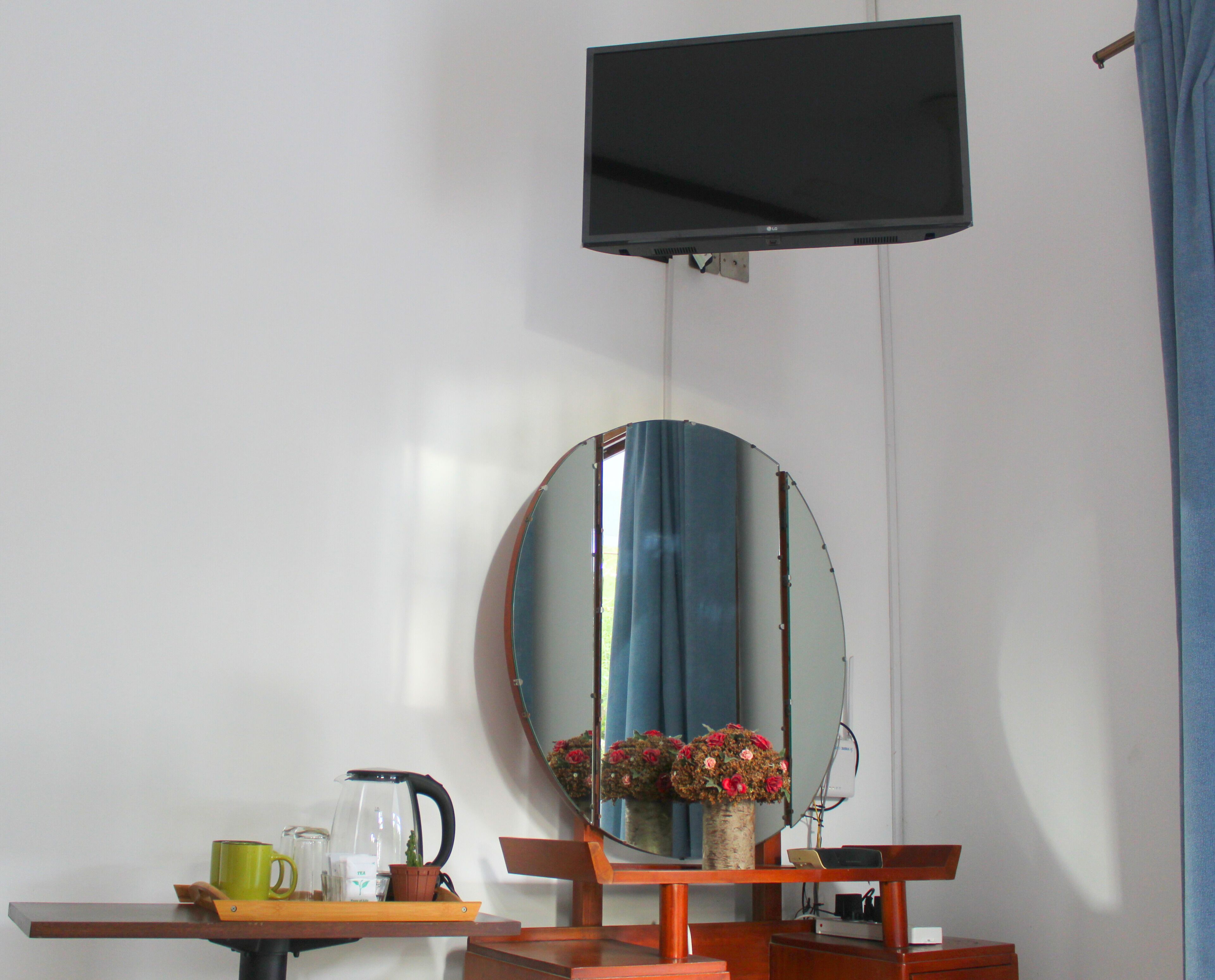 A room with a flat-screen TV mounted on the wall, a round mirror reflecting a window and a vase of flowers, and a wooden table with a kettle, mug, and a small plant.