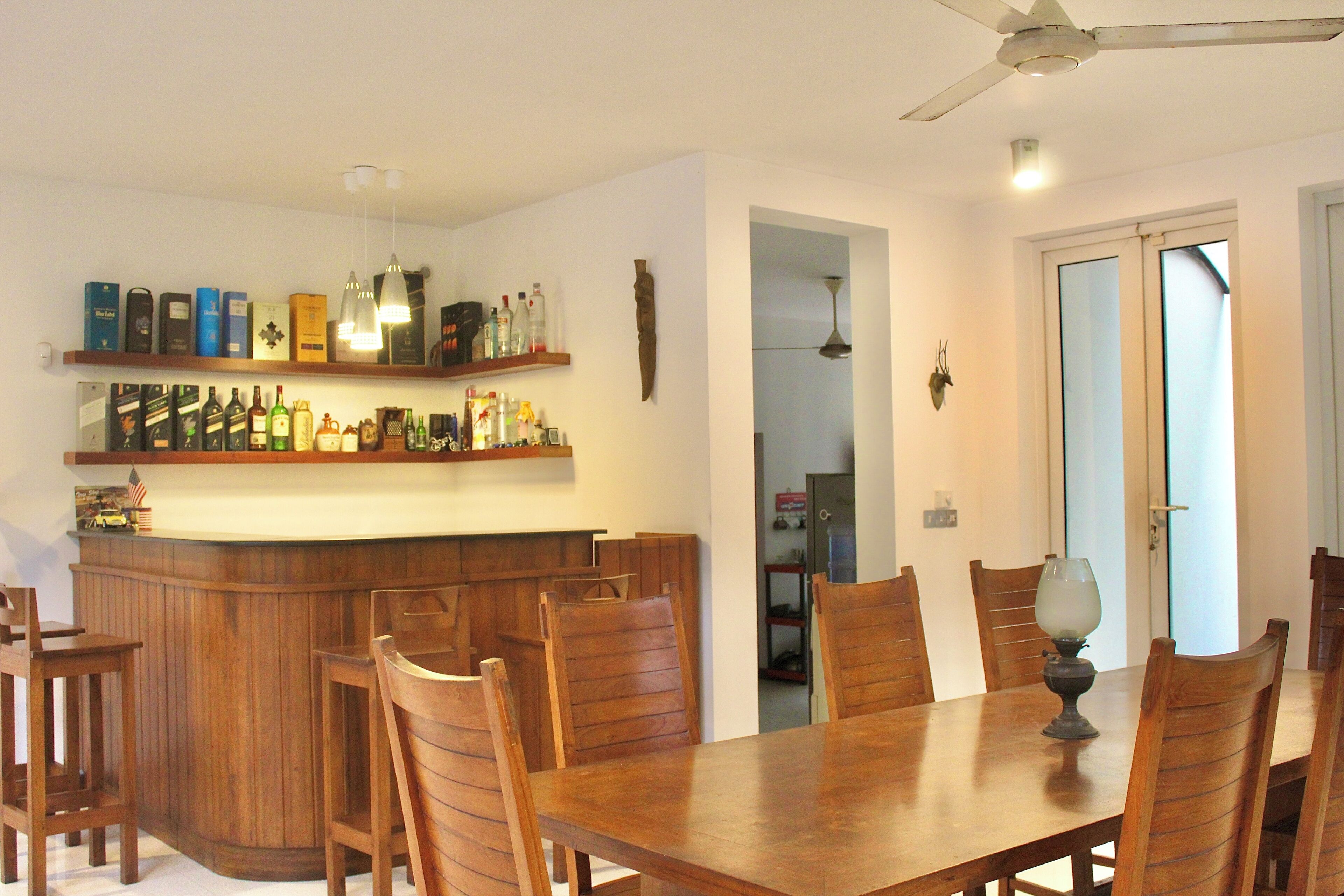 A dining area with a wooden table and chairs, a bar with shelves of bottles, and a ceiling fan.