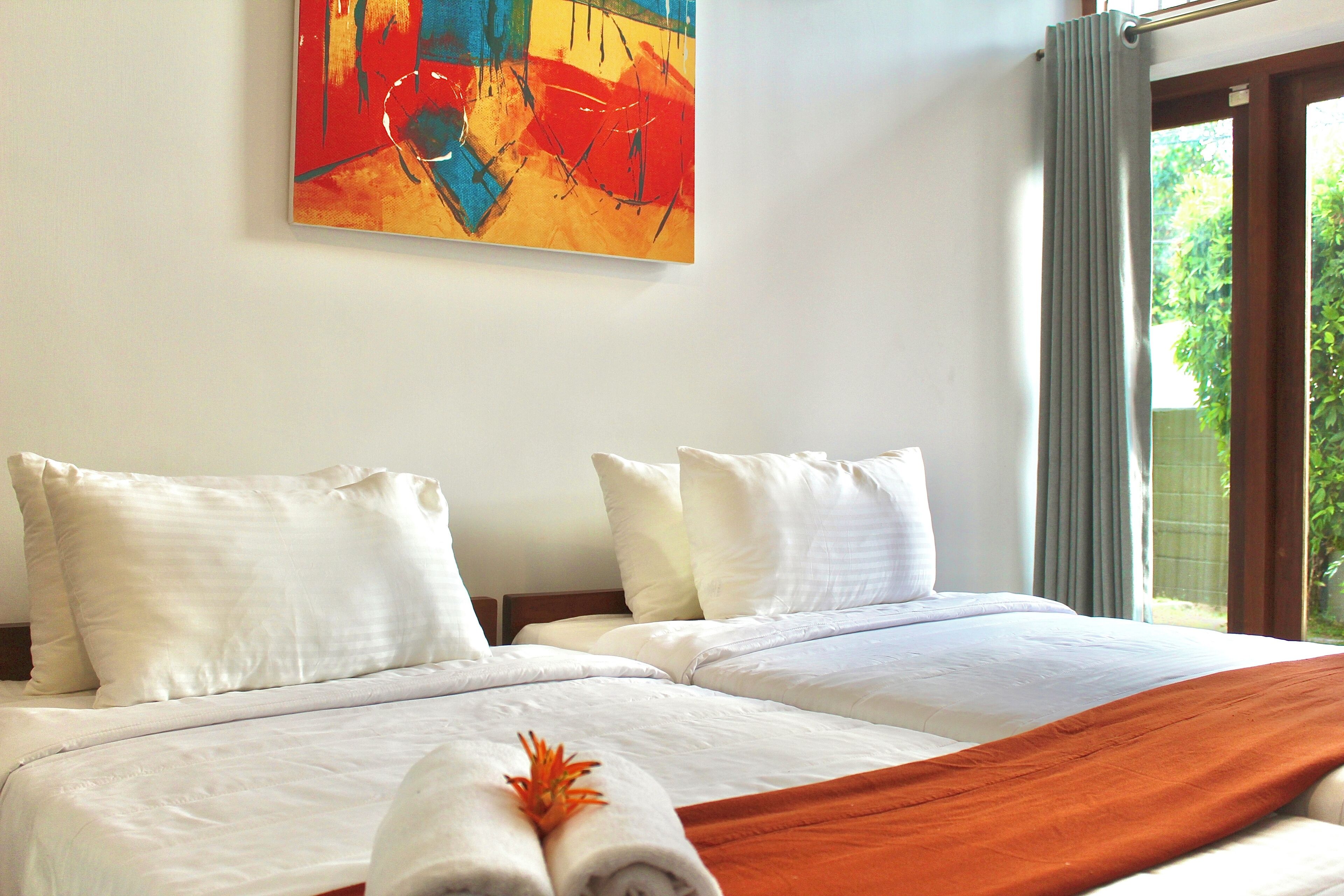 Ein Hotelzimmer mit zwei Betten, weißer Bettwäsche, einem farbenfrohen abstrakten Gemälde an der Wand und Blick auf Grün durch das Fenster.