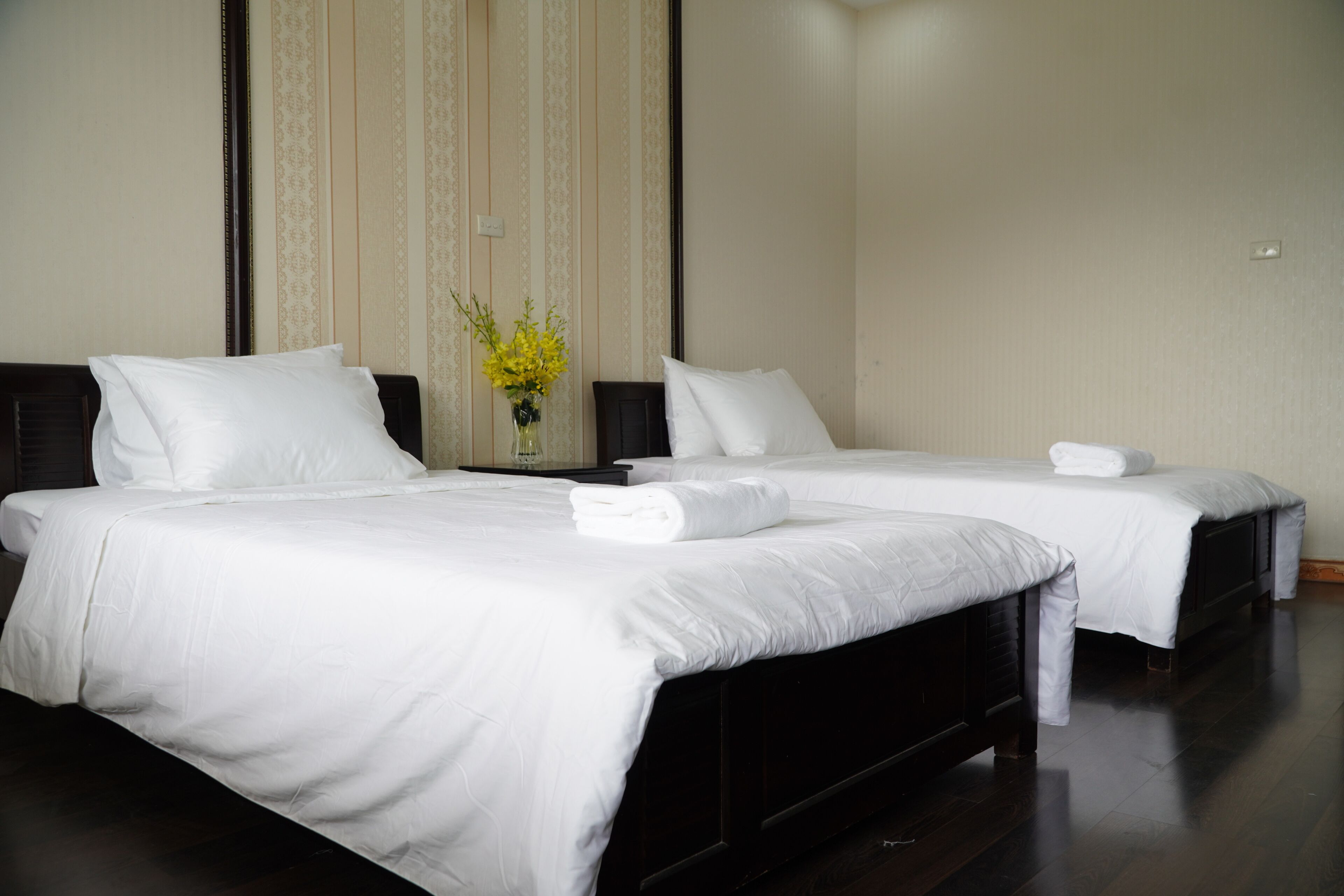 Two single beds with white linens, a dark wooden headboard, and a vase with yellow flowers on a nightstand.