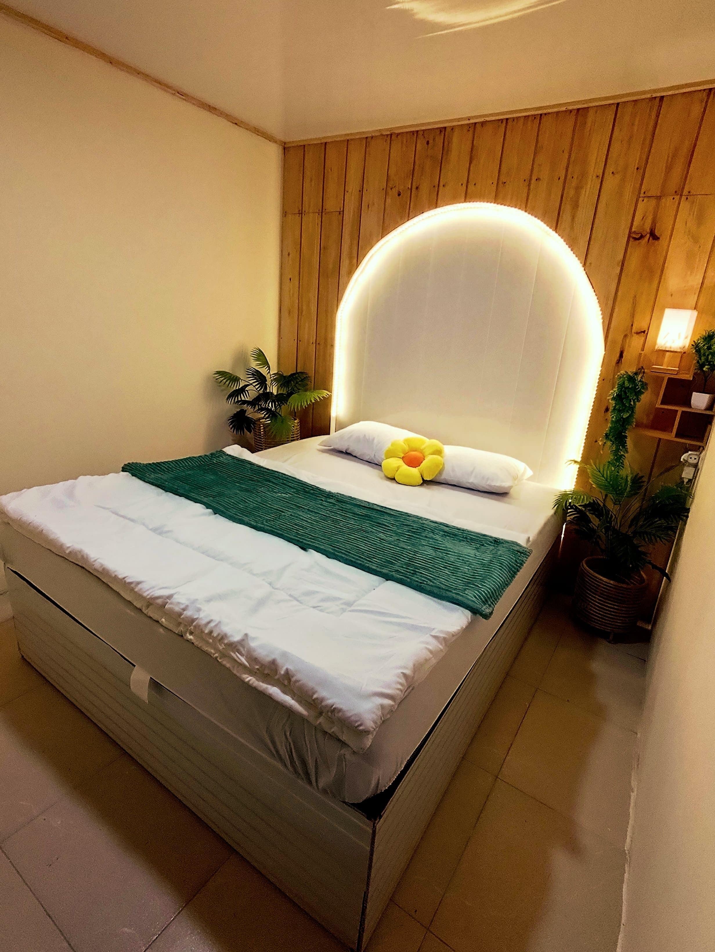 A neatly made bed with a white mattress, a green blanket, and a yellow flower pillow. There are two potted plants on either side of the bed.