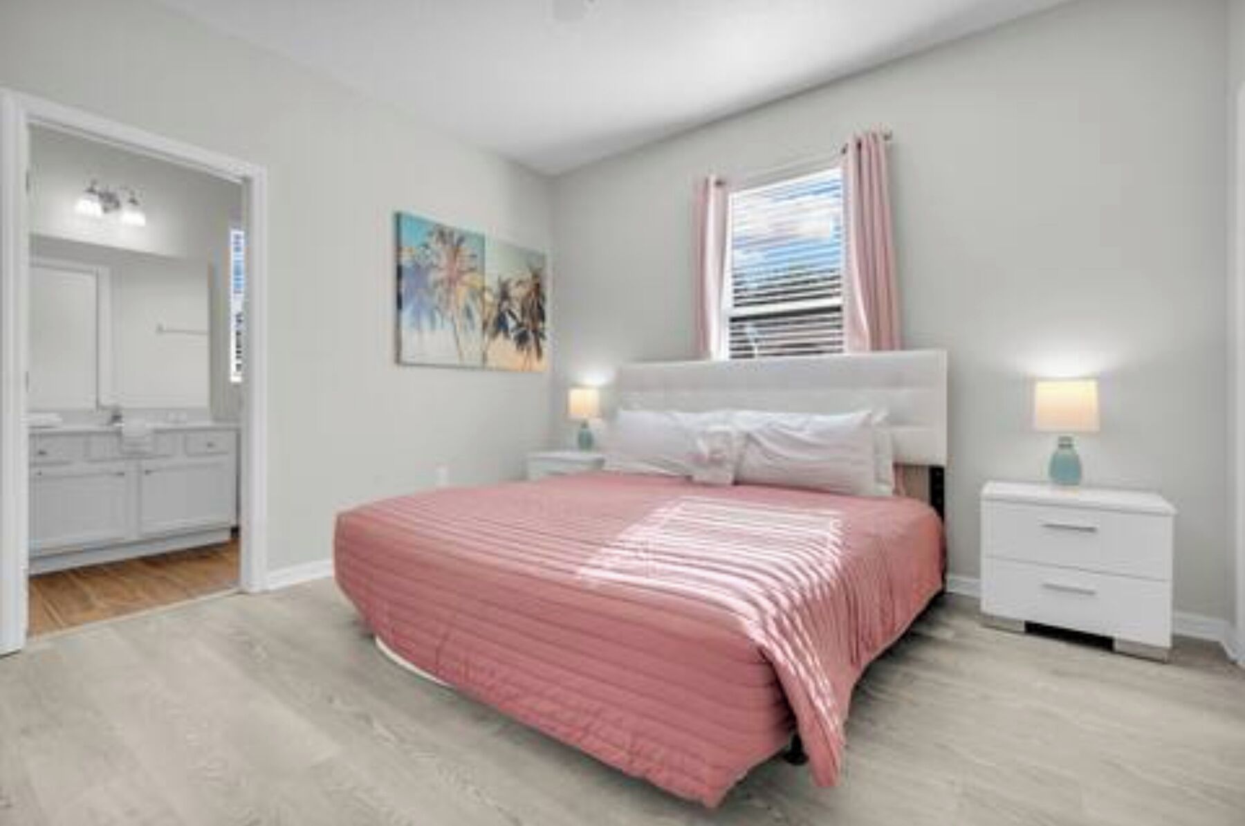 A bedroom with a bed, bedside tables, a window with curtains, a painting on the wall, and a bathroom visible through an open door.