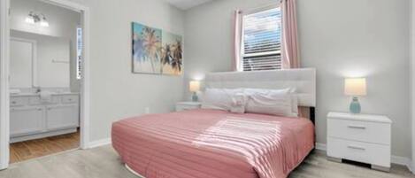 A bedroom with a bed, bedside tables, a window with curtains, a painting on the wall, and a bathroom visible through an open door.