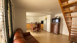 A living room with a brown leather sofa, a wooden chair, a dining table with chairs, a kitchen area with cabinets and a TV, and a wooden staircase.
