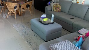 A modern living room with a grey sofa, a dining table with wicker chairs, a kitchen in the background, and a decorative clock on the wall. - Large 3x bedroom apartment directly opposite beach. Very comfortable and cosy. (Coffs Harbour)