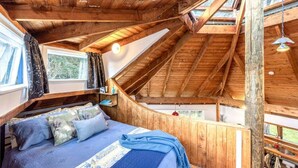 Une chambre mansardée chaleureuse avec un plafond en pente, des poutres en bois, un lit avec une literie bleue et une fenêtre avec des rideaux.