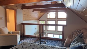 A cozy attic bedroom with a sloped ceiling, wooden beams, a large window, a bed, a chair, and a small table. - Lakefront home with private dock on Lake Ossipee in Freedom, NH sleeps up to 10 (Freedom)