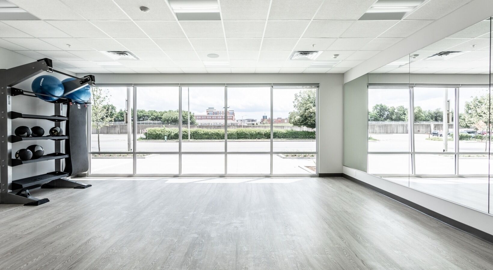 A spacious gym with large windows, a wooden floor, and exercise equipment including a squat rack and medicine balls.