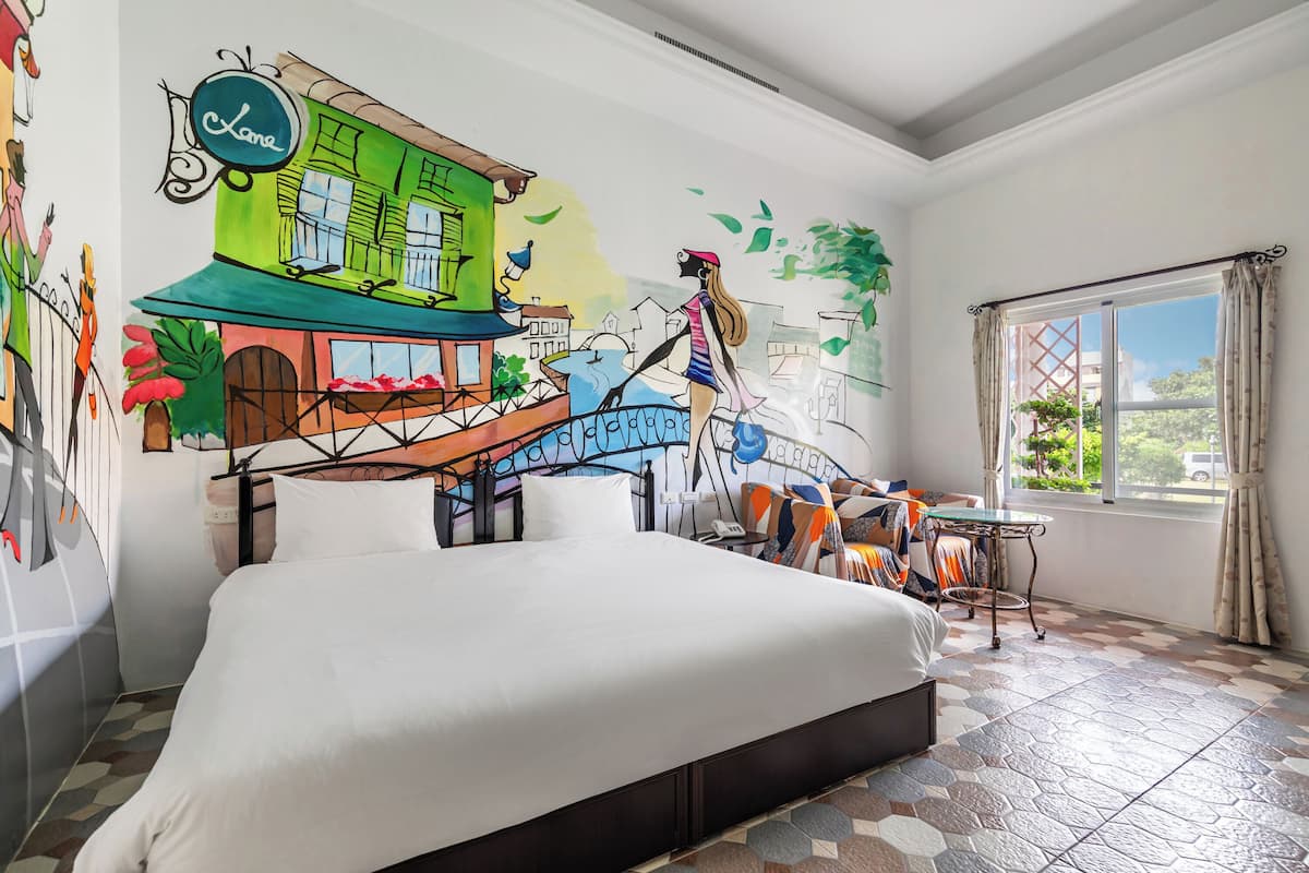 A hotel room with a large bed, a small table, a window with curtains, and a mural on the wall depicting a street scene.