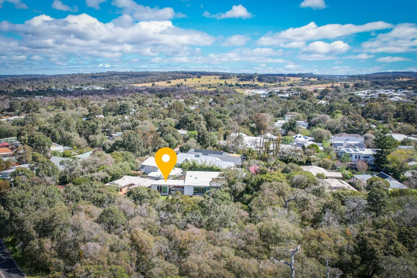 An aerial view of a residential area with houses surrounded by trees, a clear sky, and a yellow location marker.
