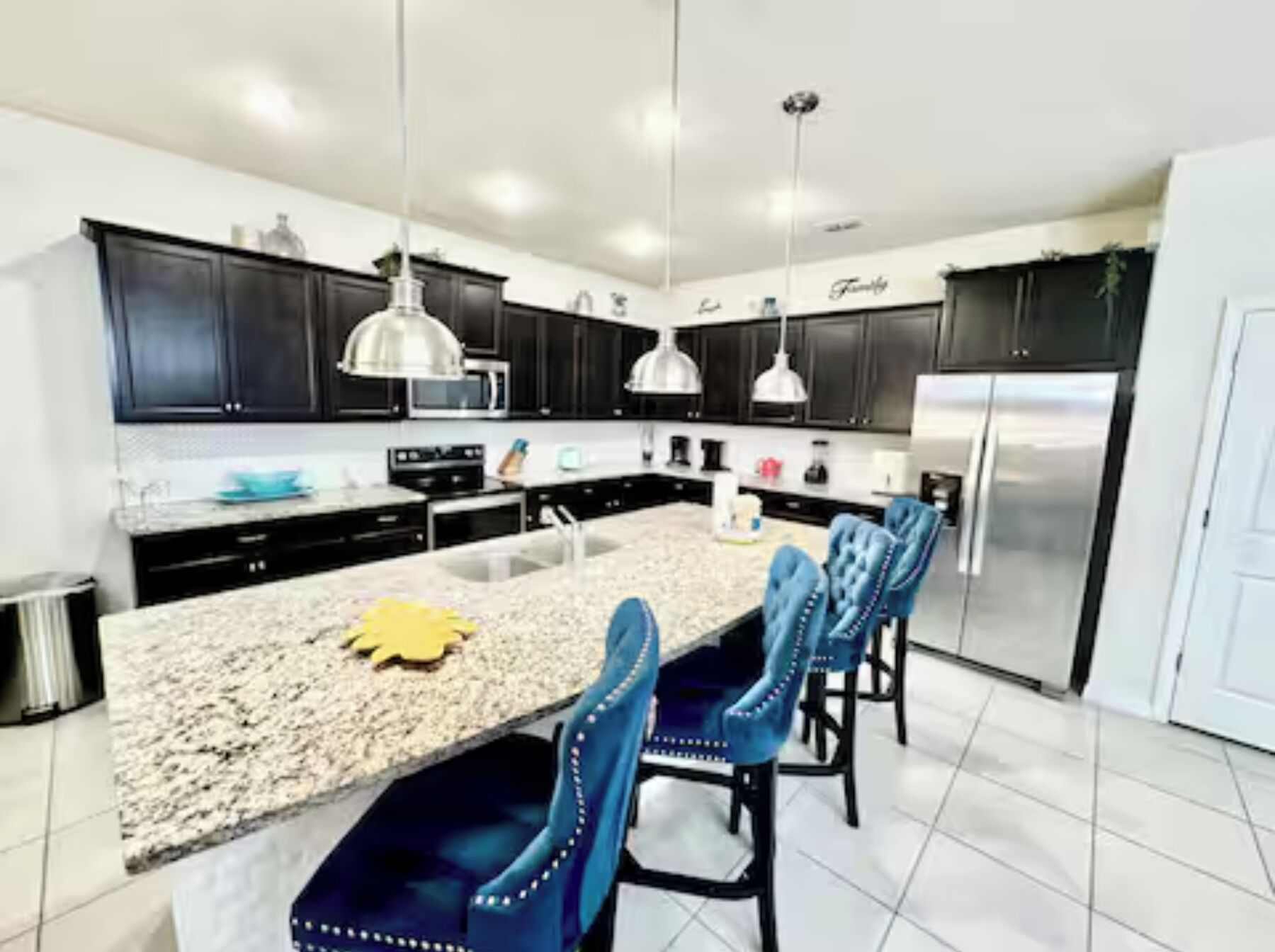 A modern kitchen with dark cabinets, a granite countertop, a stainless steel refrigerator, and blue upholstered chairs.