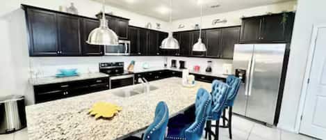 Una cocina moderna con armarios oscuros, encimera de granito, refrigerador de acero inoxidable y sillas tapizadas en azul.