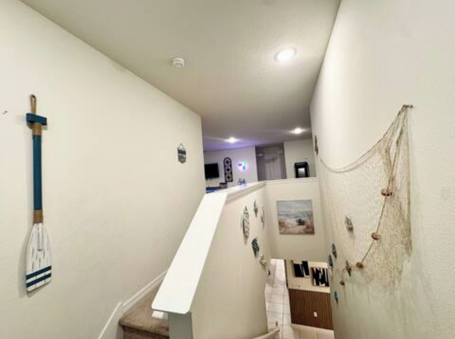 Un pasillo estrecho con una escalera blanca, un aplique de luz y un pequeño cuadro.