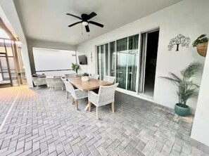 A modern dining area with a wooden table and white wicker chairs, a ceiling fan, and a large sliding glass door leading to a balcony. - The Disney Mansion by Orlando (Davenport)