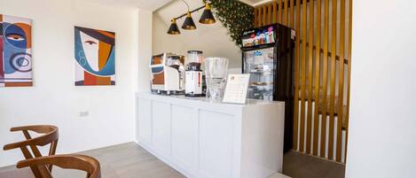 A modern hotel lobby with a reception desk, coffee machines, and a menu board. There are abstract paintings on the wall and wooden slats on the right.