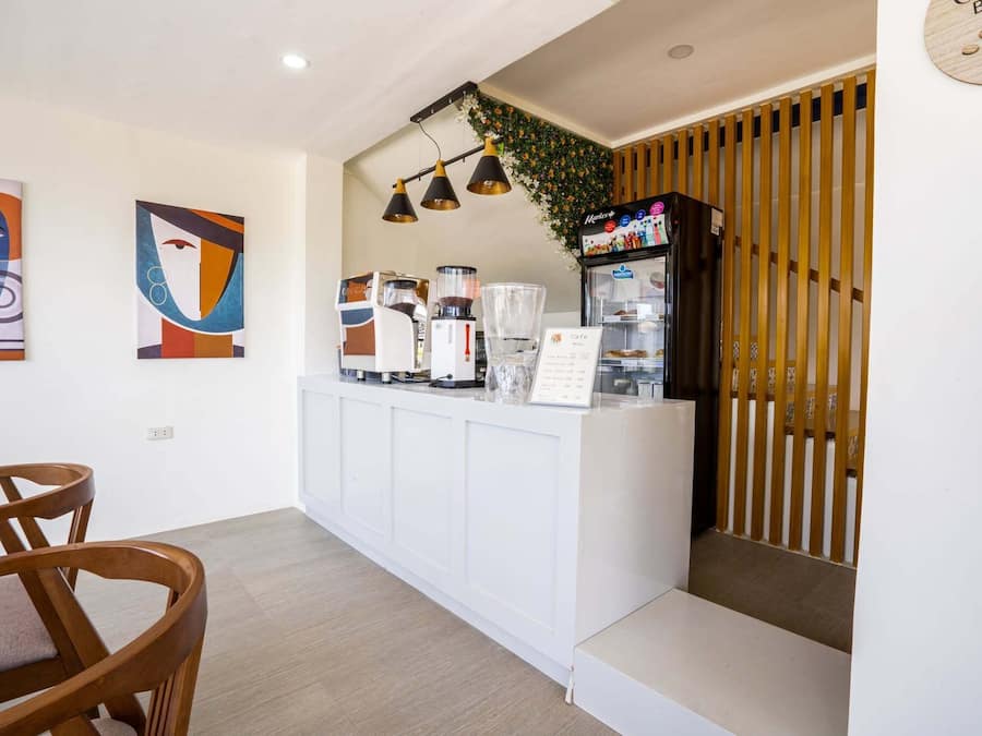 A modern hotel lobby with a reception desk, coffee machines, and a menu board. There are abstract paintings on the wall and wooden slats on the right.