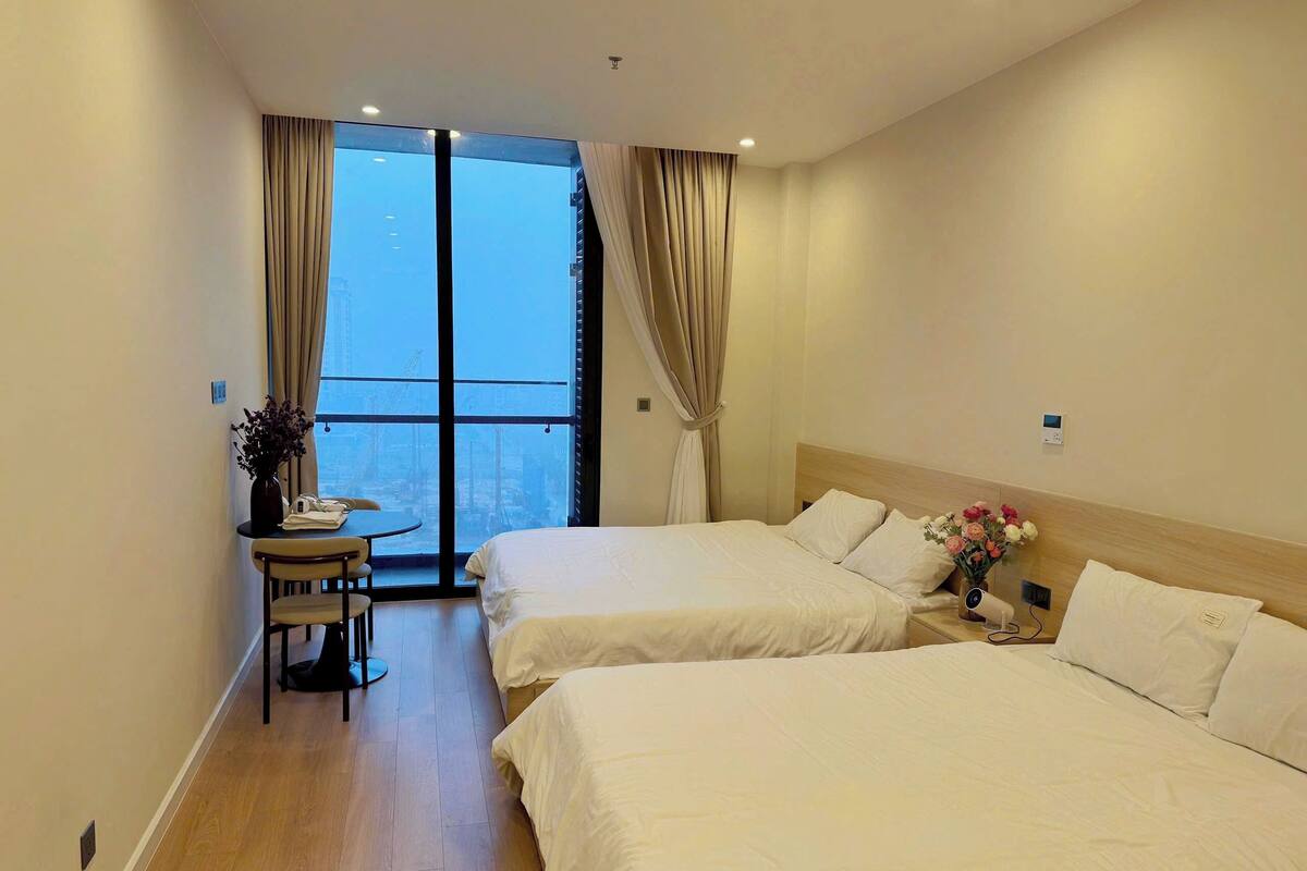 A hotel room with two beds, a small table with a chair, a vase with flowers, and a view of a cityscape through large windows.