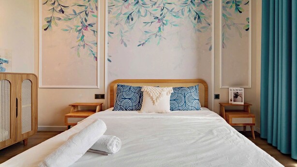 A neatly made bed with white linens, two blue patterned pillows, and a white throw blanket. There are two wooden nightstands with a small table on each, and a wooden wardrobe with a woven design. The room has a large window with blue curtains, and a wall with a floral painting. - Halong Seaside Retreat (Bai Chay)
