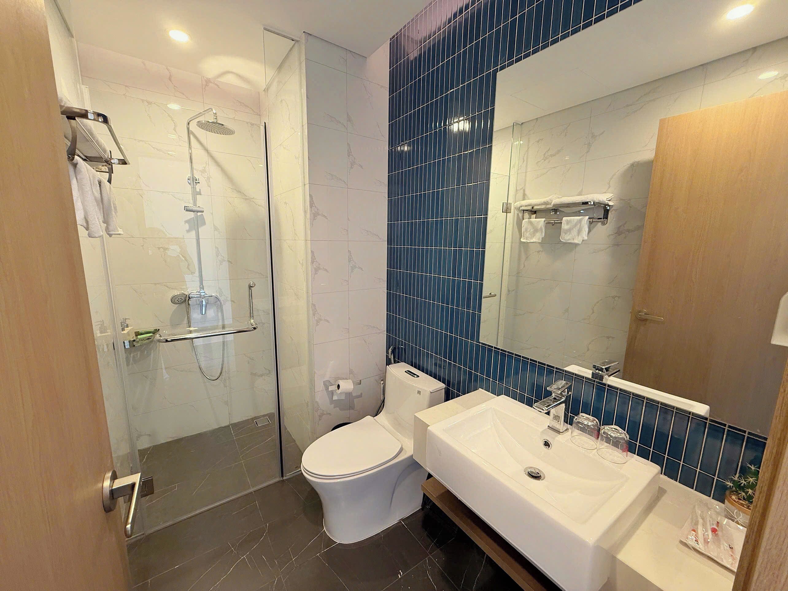 Un baño moderno con ducha envuelta en vidrio, inodoro blanco, lavabo grande y espejo con detalles de azulejos azules.