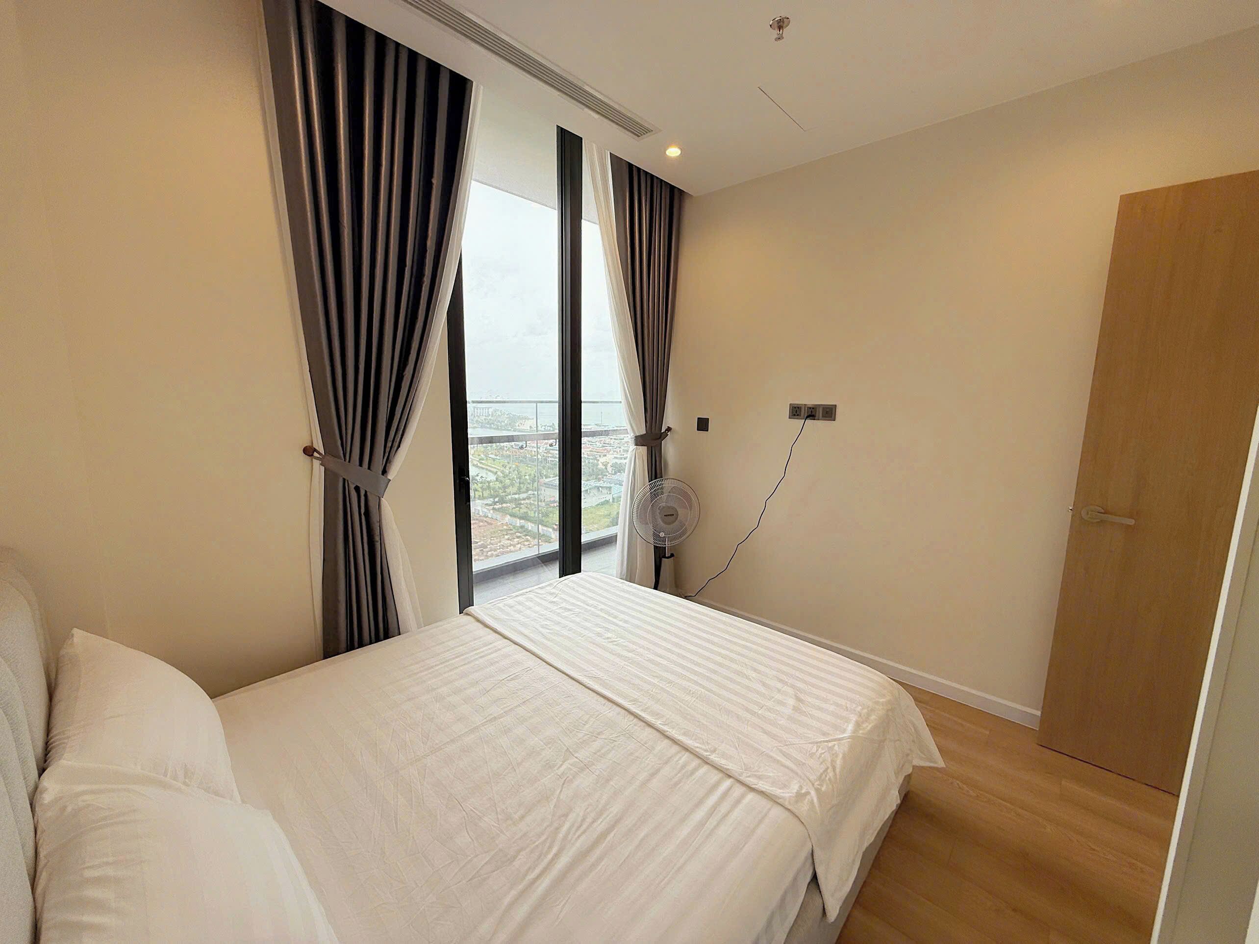 A bedroom with a large bed, a window with curtains, a wall-mounted fan, and a wooden door.