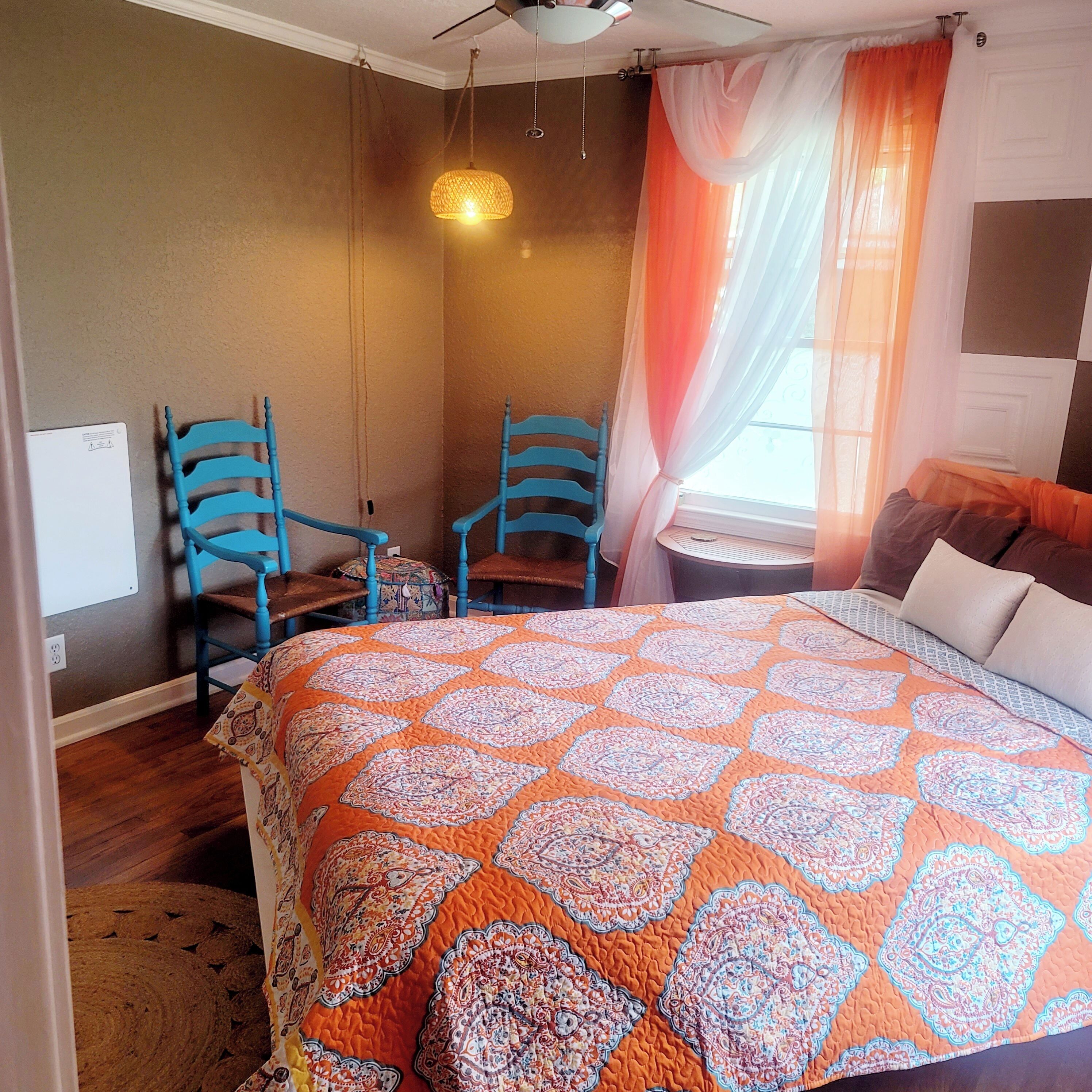 Ein Schlafzimmer mit einem großen Bett, zwei blauen Stühlen, einem kleinen Tisch und einem Fenster mit durchsichtigen Vorhängen.