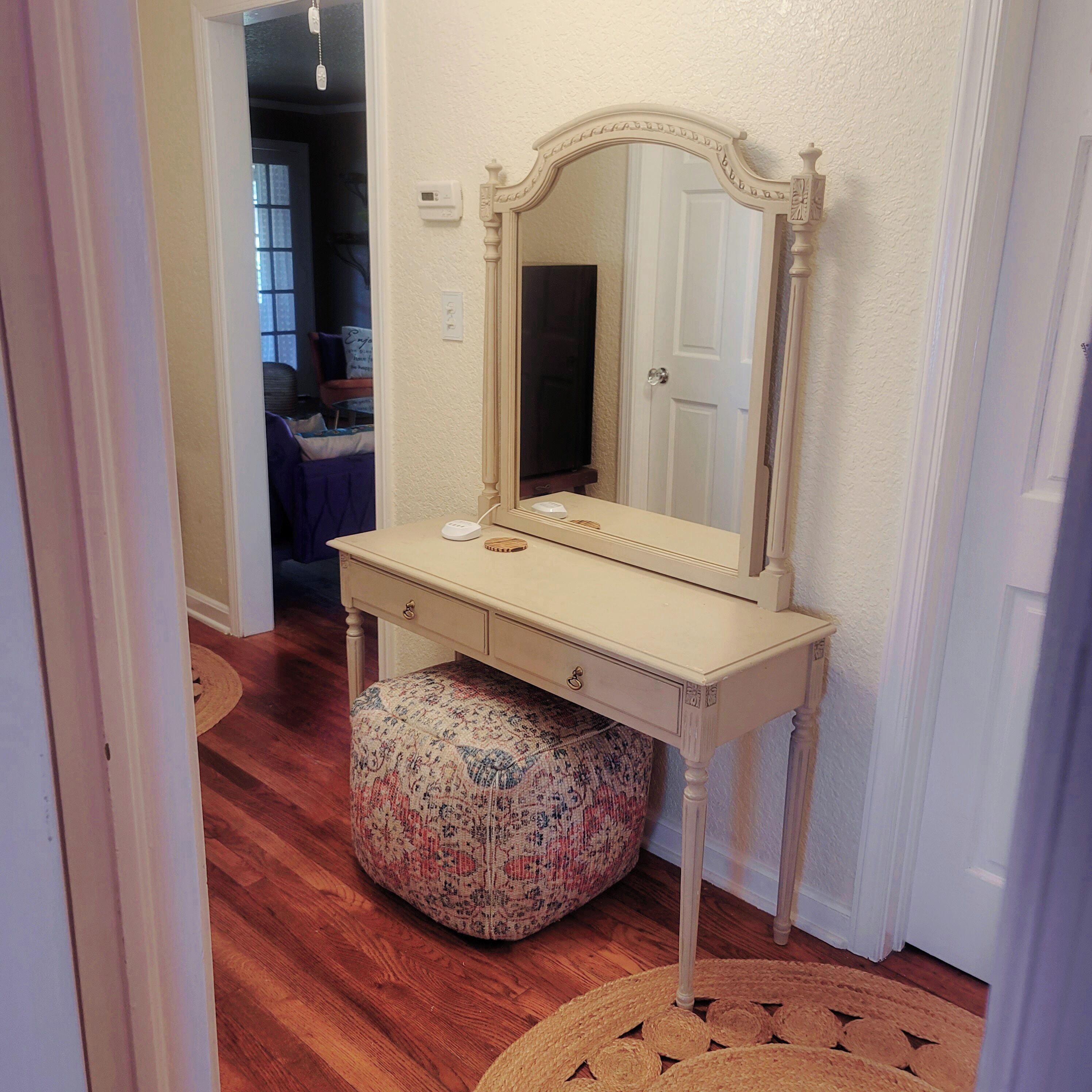 A small, cream-colored vanity with a mirror, a drawer, and a round ottoman with a floral pattern.