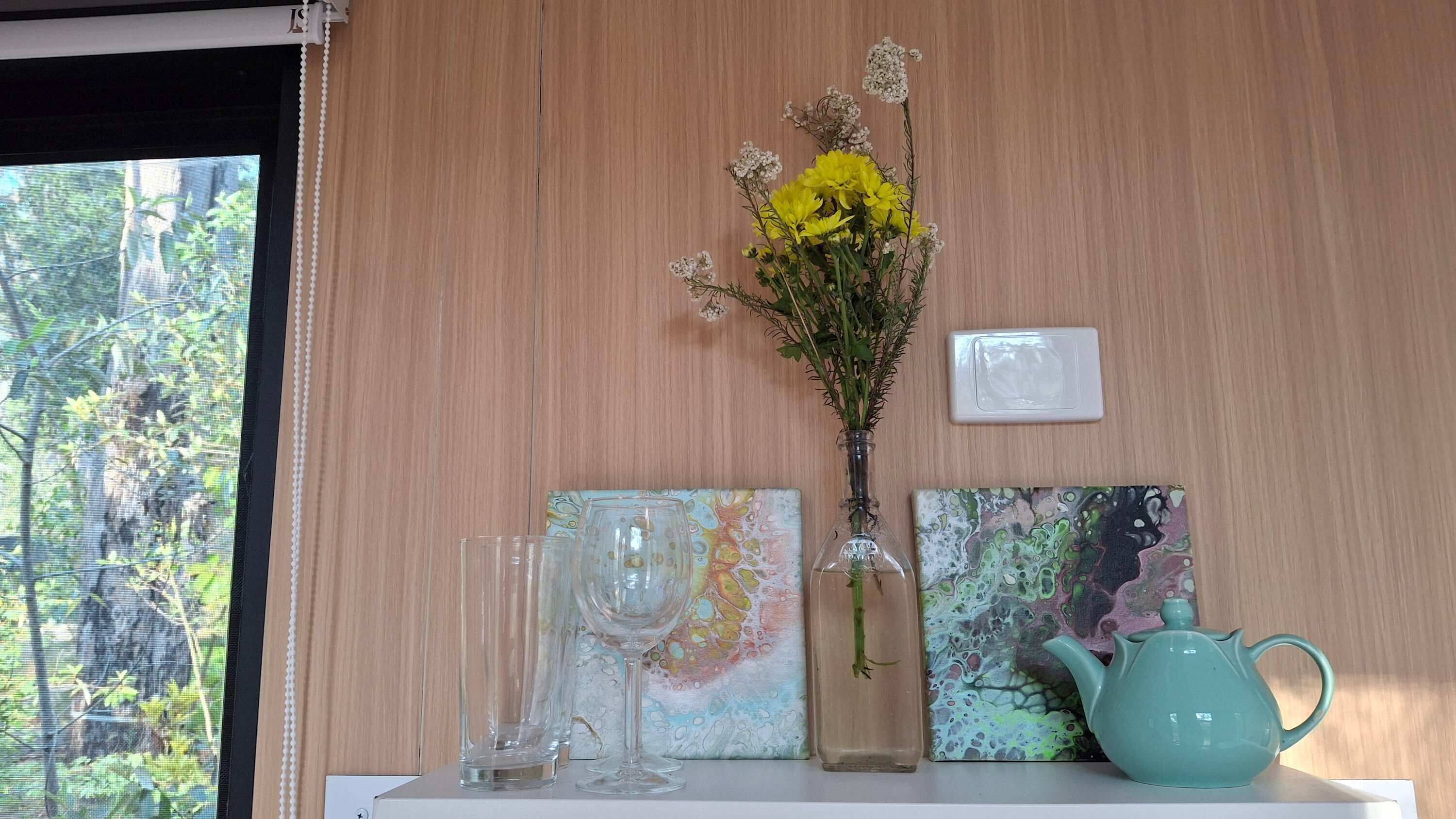 Une étagère avec un vase rempli de fleurs jaunes, un vase en verre, une théière et deux tableaux abstraits.