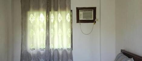 Un dormitorio con una cama individual, una ventana con cortinas, un equipo de aire acondicionado instalado en la pared y una mesita de noche con lámpara.