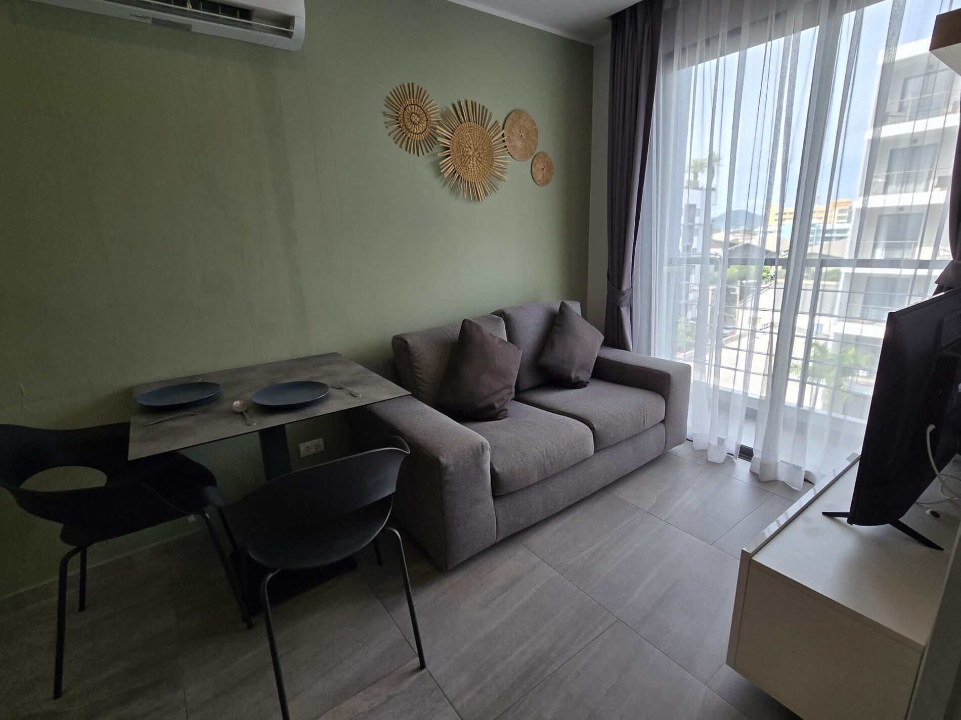 A modern living room with a grey sofa, a dining table with chairs, and a large window with sheer curtains.