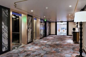 Un couloir d’hôtel avec un tapis à motifs, des portes d’ascenseur et une lampe.