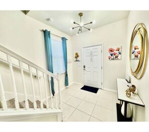 A clean, well-lit hallway with a white staircase, a door with a doormat, and a wall-mounted mirror. - The Dream Getaway Retreat Near Disney (Davenport)