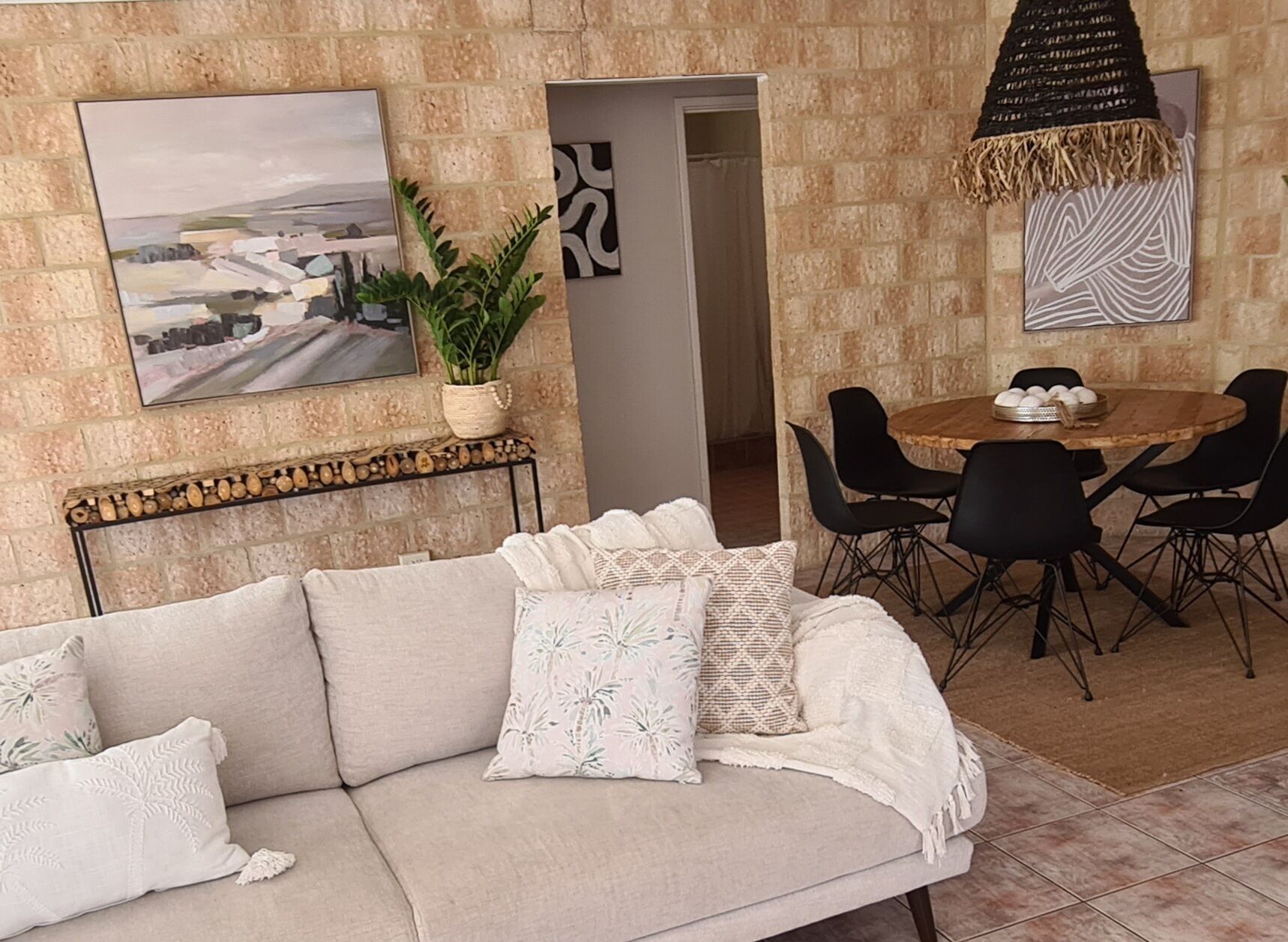 A living room with a beige sofa, a dining table with black chairs, a potted plant, and artwork on the walls.