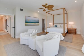 A bedroom with a four-poster bed, a ceiling fan, a painting of a beach scene, and a white armchair. - Endless Blue: Luxury Cudjoe Key Waterfront Retreat | Heated Pool, Dock, Guest House & Dog-Friendly (Cudjoe Key)