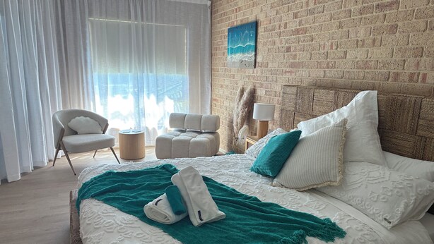 A neatly arranged hotel room with a large bed, two chairs, a small table, and a brick wall with a painting.