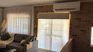 A modern living room with a grey sofa, a large window with sheer curtains, and an air conditioning unit mounted on a brick wall.