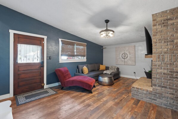 A modern living room with a wooden floor, a red chair, a grey sofa, and a brick accent wall. - Downtown Home-Heated Plunge Pool Outdoor Oasis (Loveland)