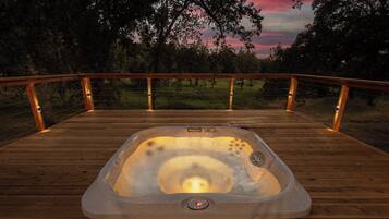 A hot tub on a wooden deck with a railing, surrounded by trees and a sunset.