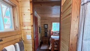 A wooden cabin interior with a hallway leading to a bedroom, a bathroom with a shower, and a small table with a lamp. - Cowboy Cabin -tucked into the woods near Hatcher Pass Recreation Area. (Palmer)