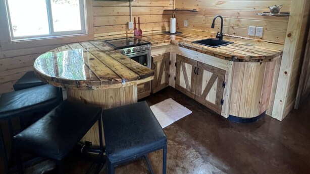 A wooden cabin interior with a kitchen area featuring a round table, bar stools, a sink, and a stove. - Cowboy Cabin -tucked into the woods near Hatcher Pass Recreation Area. (Palmer)
