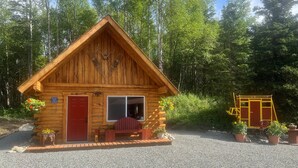A wooden cabin with a red door and windows, surrounded by gravel and greenery. - Cowboy Cabin -tucked into the woods near Hatcher Pass Recreation Area. (Palmer)