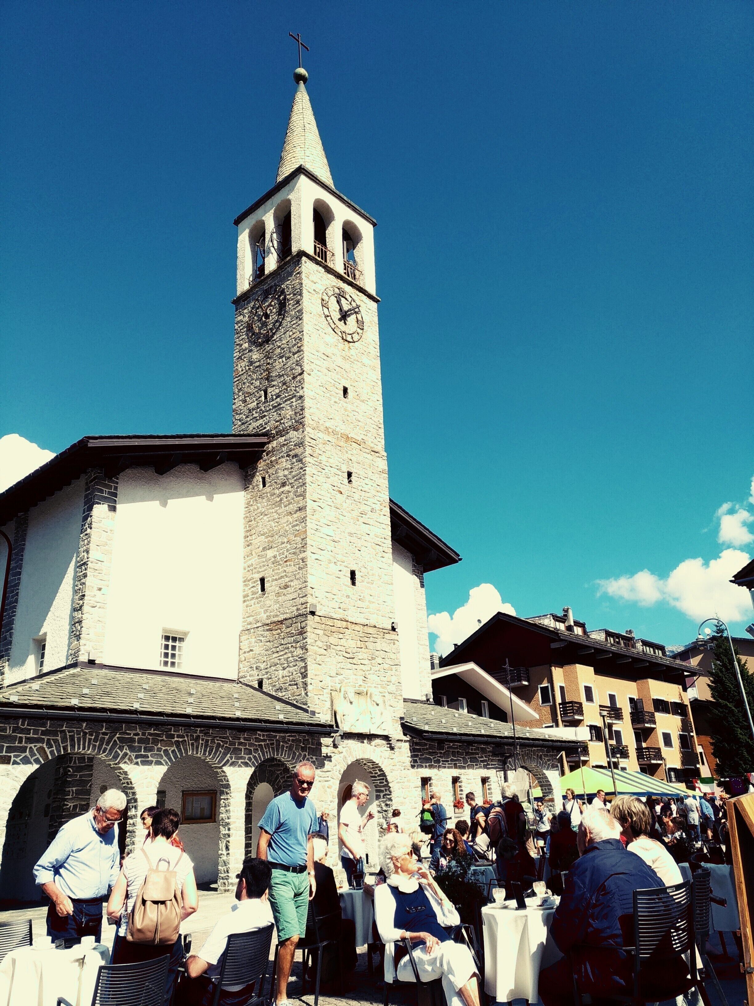 Une église historique dotée d’un clocher, d’un espace extérieur aménagé pour s’asseoir et où se trouve une foule de personnes.