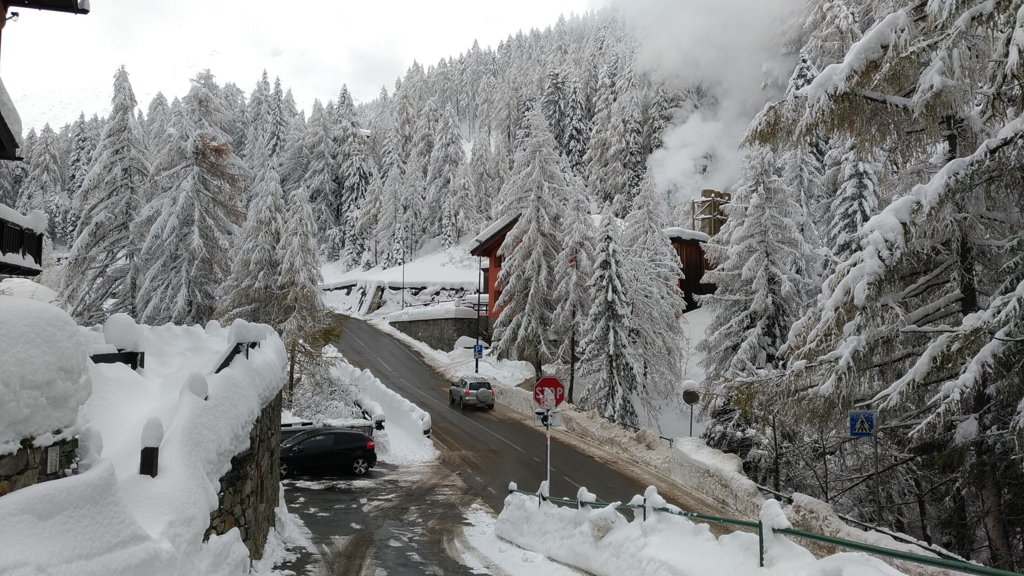 A snow-covered road with a car parked on the side, surrounded by snow-laden trees and a building with a red roof.
