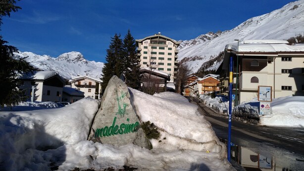 A snow-covered mountain town with a large rock inscribed with "made simmo" in the foreground, a road with a blue sign, and a hotel with multiple floors and balconies.