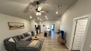 A modern living room with a grey sectional sofa, a wooden coffee table, and a TV on a wooden stand. A ceiling fan and recessed lighting are visible.